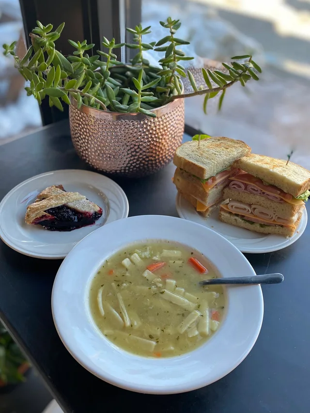 A bowl of chicken noodle soup with a separate sandwich and slice of pie.