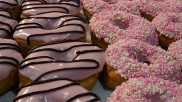 Pink-iced donuts with dark drizzle beside ring donuts covered in pink crumbs.