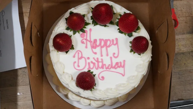 A white birthday cake with pink lettering and whole strawberries around the top.
